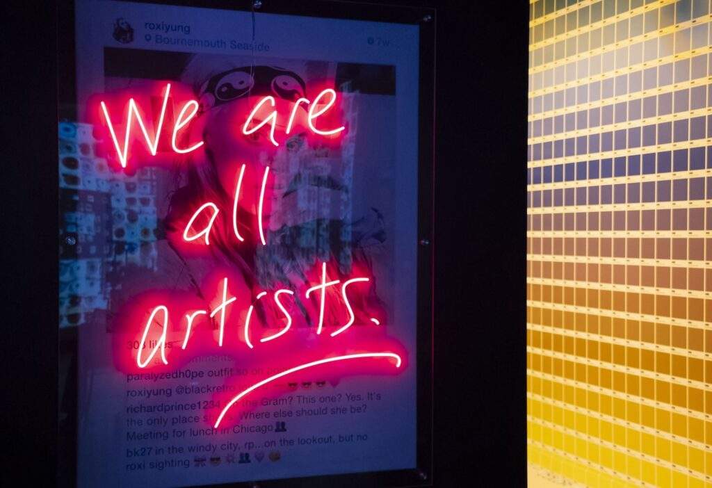 "We Are All Artists" is written in neon over a blown-up Instagram post.